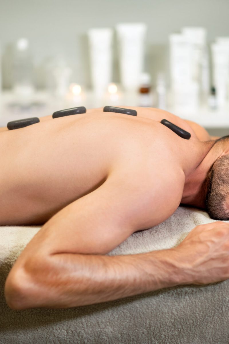 Side view of shirtless young male with towel on hips and hot stones along spines lying with closed eyes on table during massage session in spa salon